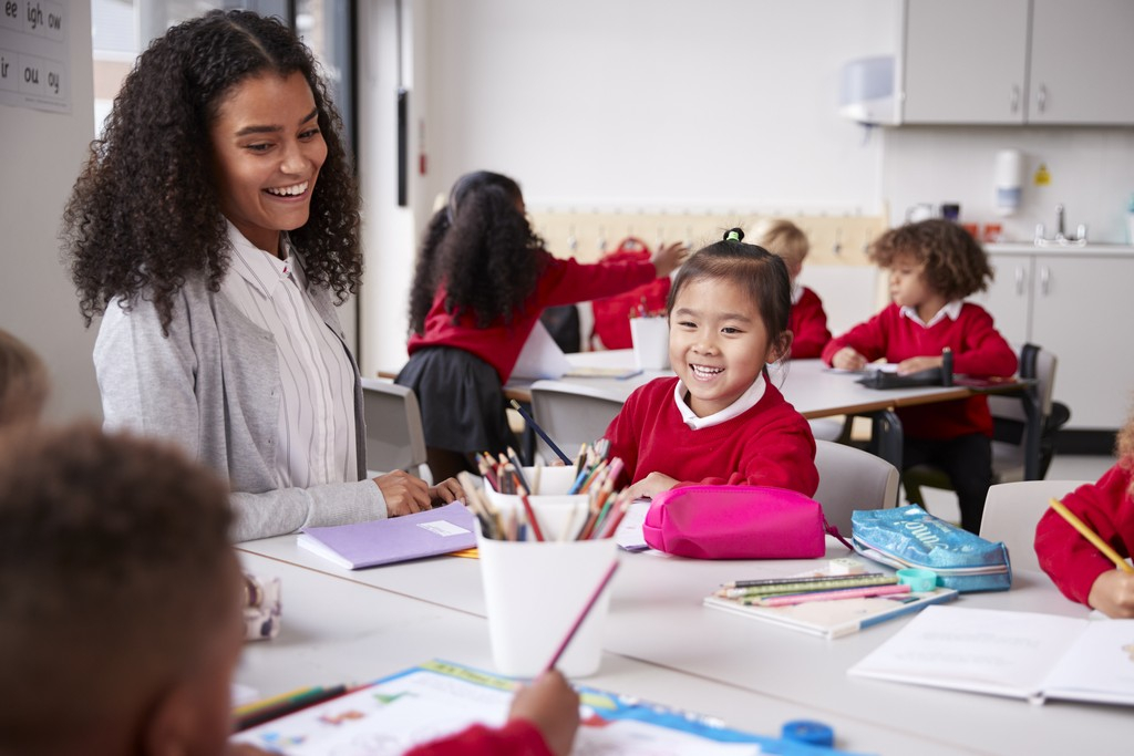 teacher and schoolgirl sitting at a table in an infant school class smiling to other kids, selective focus